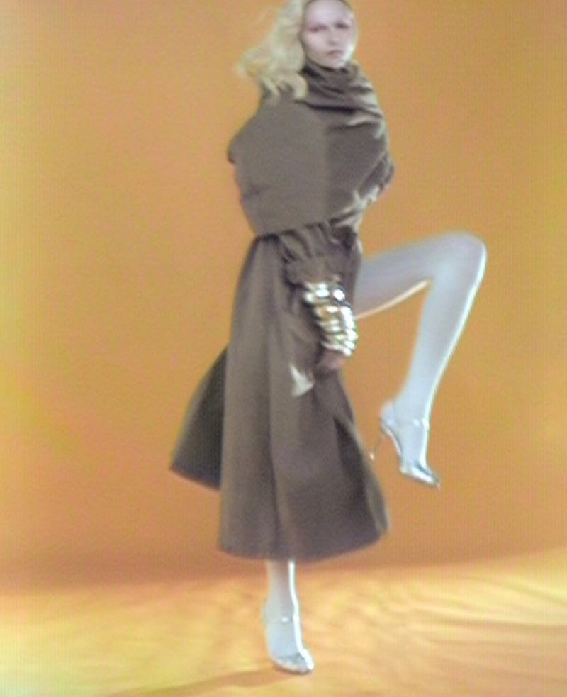 Woman in a brown coat and silver heels posing against an orange background, showcasing fashion and style in a studio setting.