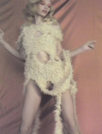 Blonde woman in textural garment poses in front of a peach-colored backdrop with diffused lighting, creating a retro, fashion-forward aesthetic.