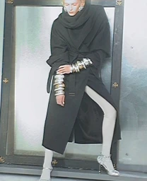 Alex Consani in a black belted coat with stacked silver bracelets standing in front of a dark framed glass door for Acne Paper