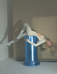 Model Alex Consani draped over a bright blue pillar wearing a white bodysuit and silver heels against a neutral studio background