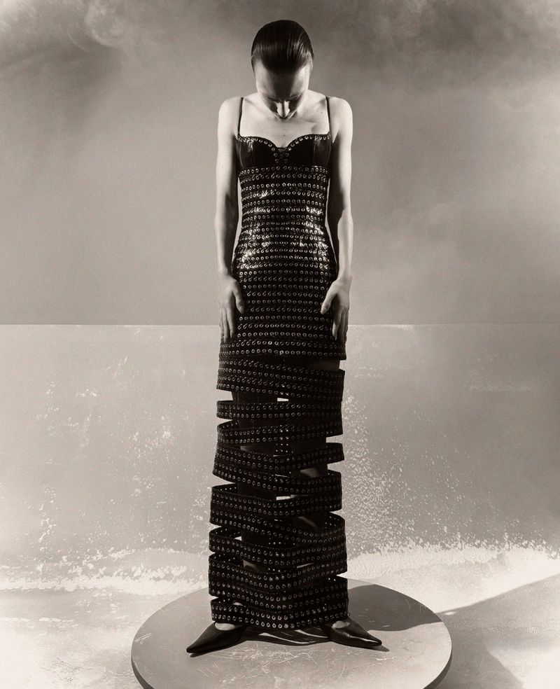 Woman in a floor-length, open-work dress stands on a platform in a studio setting with smokey lighting and a neutral palette.