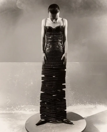 Woman in a floor-length, open-work dress stands on a platform in a studio setting with smokey lighting and a neutral palette.