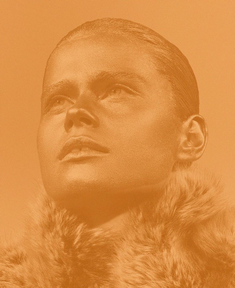 Woman with metallic gold body paint and fur collar poses in a close-up portrait with a neutral expression against a solid gold background.