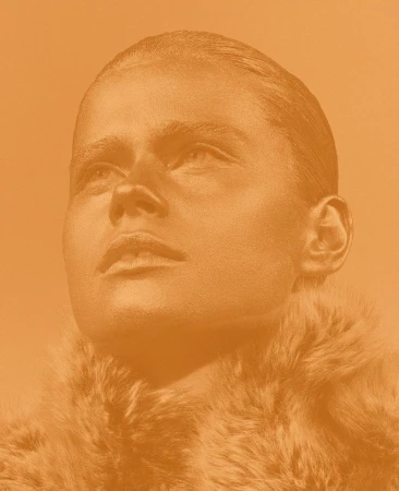 Woman with metallic gold body paint and fur collar poses in a close-up portrait with a neutral expression against a solid gold background.