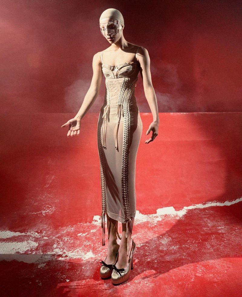 Fashion model posing in studio wearing a dress with corset, matching head covering, platform heels, and a neutral color palette.