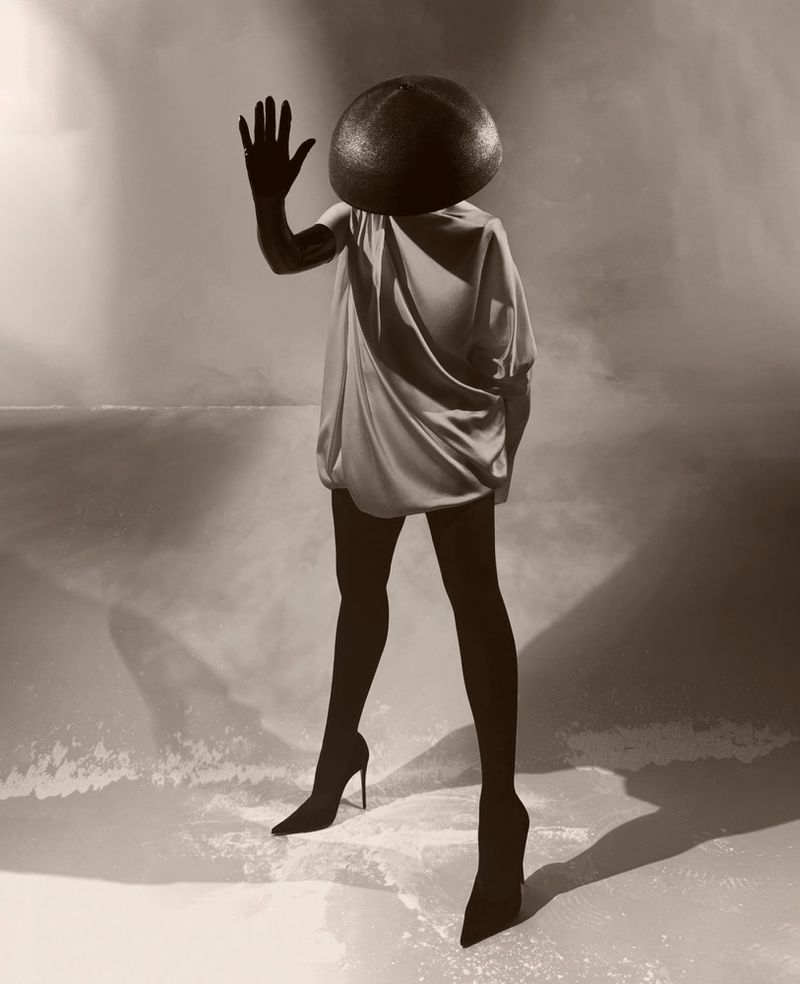 Person in stylish dress, black tights, and heels poses with hand raised in a monochrome, stylized setting with a spherical head covering.