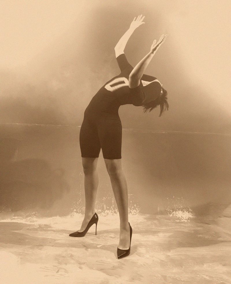 Sepia image of a woman in sports clothing and high heels, bending backward with arms raised against a blended backdrop of land and sky.