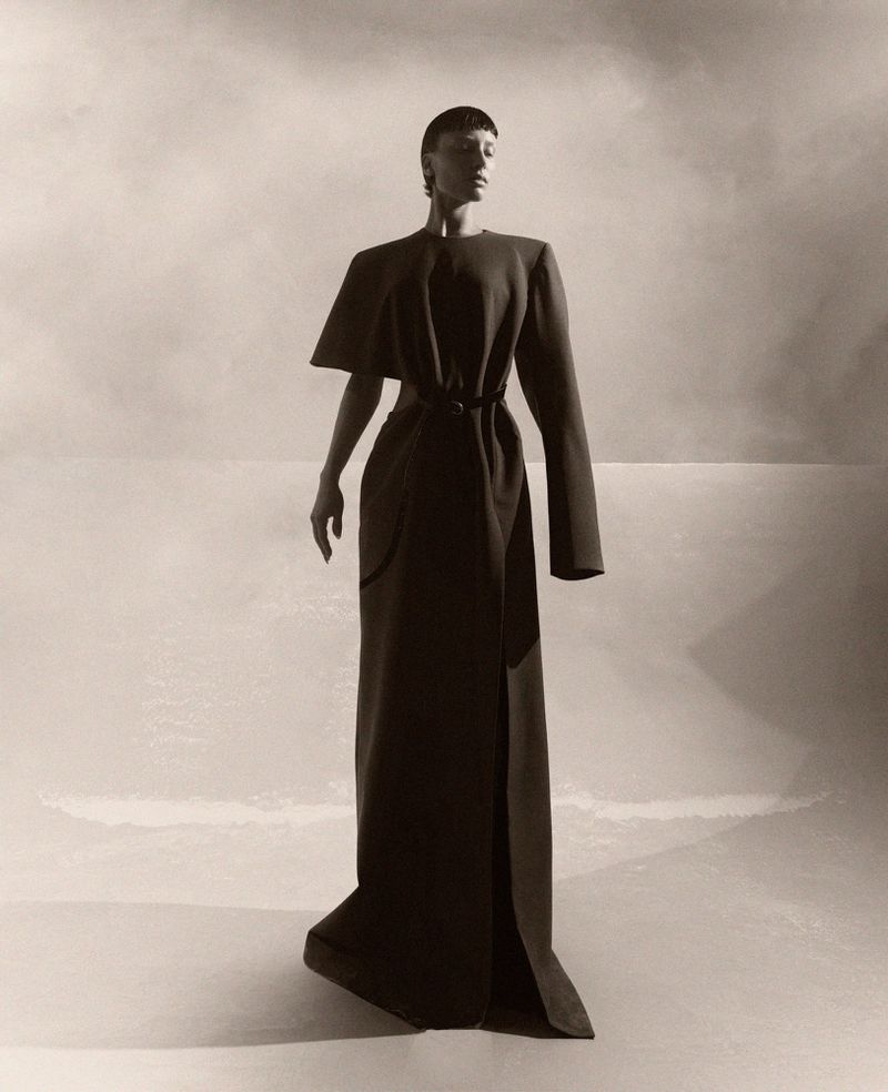 A woman stands in a long, elegant dark dress, featuring unique shoulder details and a tied waist, set against a neutral background.
