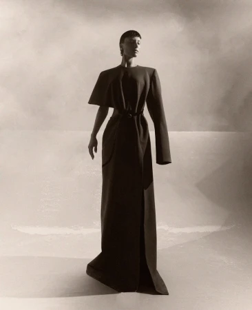 A woman stands in a long, elegant dark dress, featuring unique shoulder details and a tied waist, set against a neutral background.