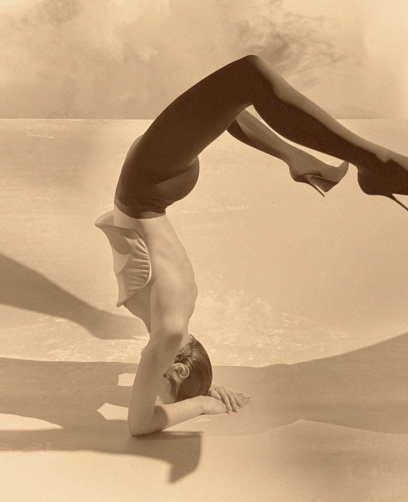A woman in a light-colored bathing suit and high heels performs a headstand on a light surface with a light background.