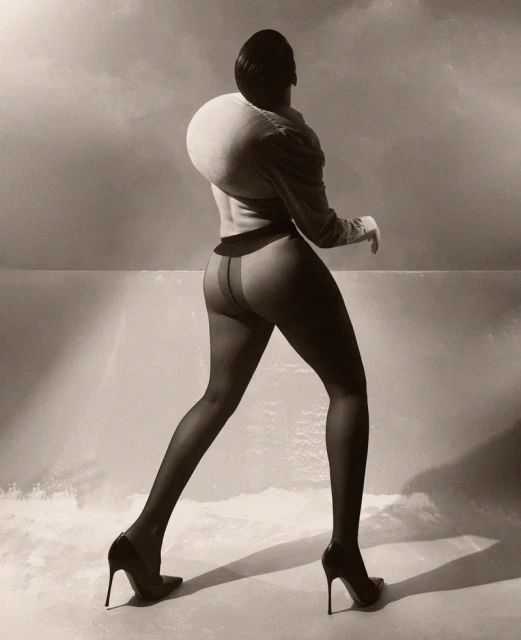 Woman in a sculptural top and stockings walks in front of a cloudy sky. She wears high heels in a sepia-toned, fashion-style photograph.