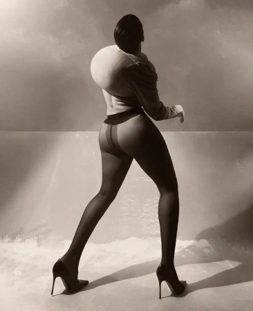 Woman in a sculptural top and stockings walks in front of a cloudy sky. She wears high heels in a sepia-toned, fashion-style photograph.