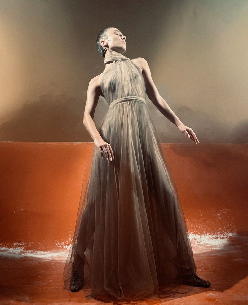 Woman in flowing gown strikes a pose in a photo studio with warm lighting, orange wall, and bright floor creating dramatic contrast.