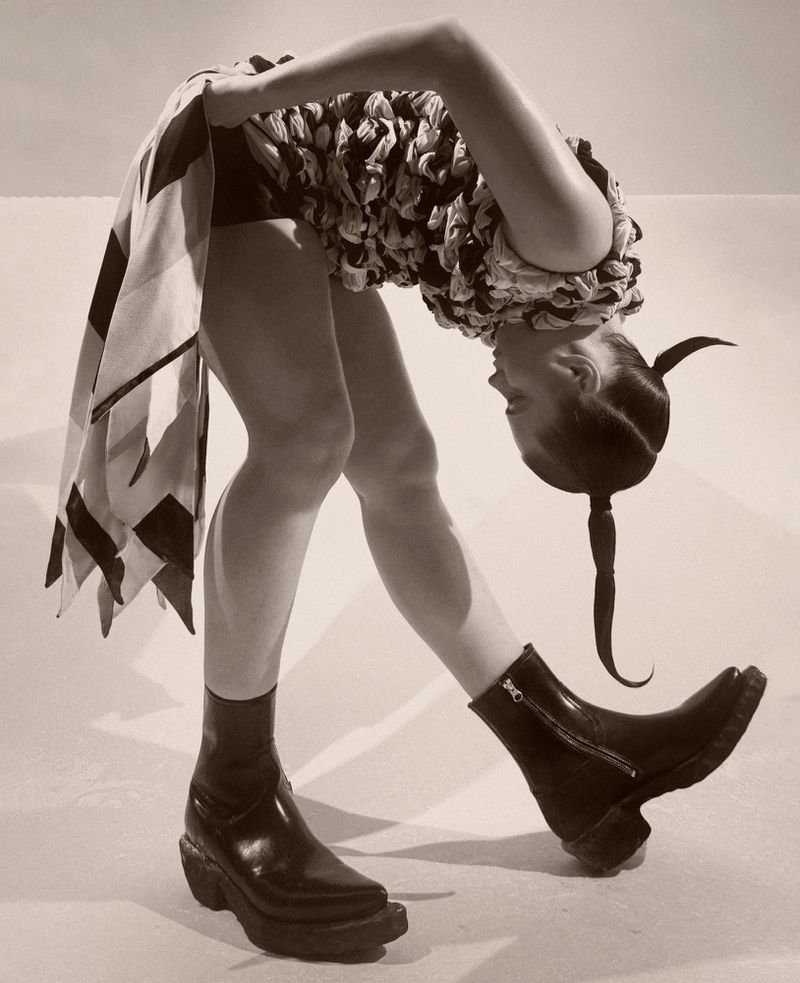 Woman in stylish black leather platform boots and draped fabric top posing in a studio with a white backdrop, artistic fashion shot.
