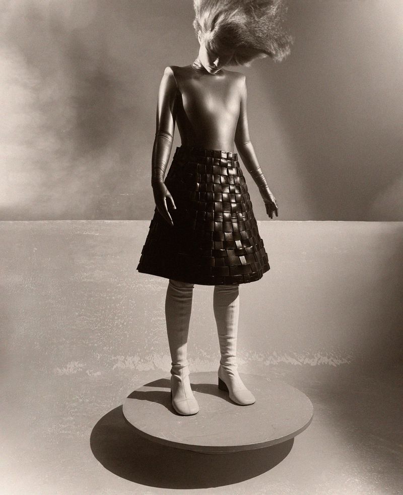 Fashion shot of a woman in metallic bodysuit, woven skirt, and boots, standing on a circular platform against a neutral background.
