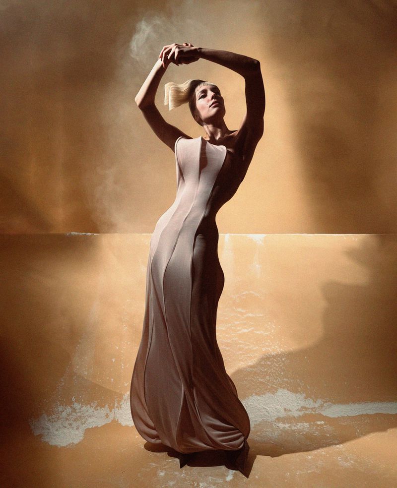 Woman poses in a flowing beige gown against a textured orange background, hands raised, creating a dramatic and stylish fashion image.