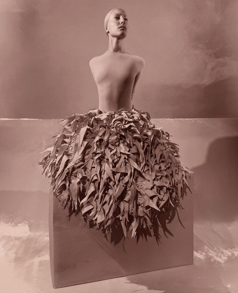 Sepia-toned fashion shot featuring a model atop a cube, wearing a skirt of layered leaves against a backdrop of clouds and a horizon.
