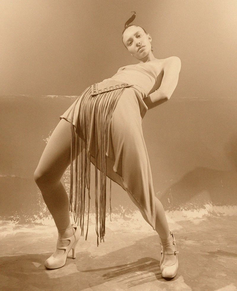 Fashionable woman strikes a pose in a beige dress with fringe and high heels against a neutral backdrop, showcasing an editorial style.
