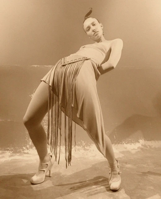 Fashionable woman strikes a pose in a beige dress with fringe and high heels against a neutral backdrop, showcasing an editorial style.