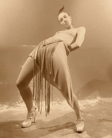 Fashionable woman strikes a pose in a beige dress with fringe and high heels against a neutral backdrop, showcasing an editorial style.