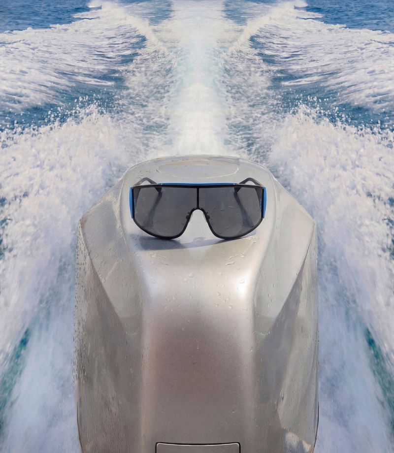Sunglasses rest on a boat's silver engine cover against a backdrop of blue water and foamy wakes under bright, natural lighting.