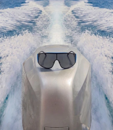 Sunglasses rest on a boat's silver engine cover against a backdrop of blue water and foamy wakes under bright, natural lighting.