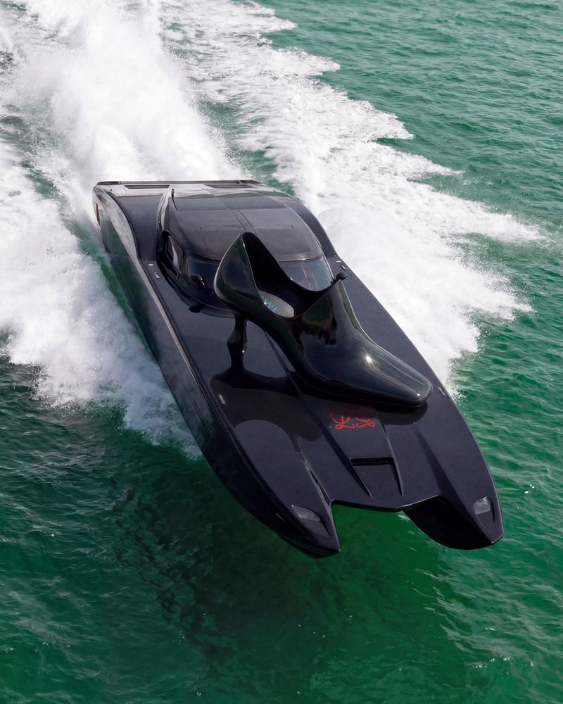 A glossy black speedboat speeding on the green water, with a shoe-shaped decor on the boat. White foamy water trails behind the boat.
