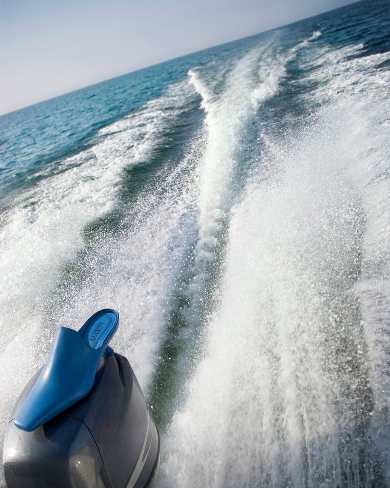 A blue high-heeled shoe rests on a boat motor against a background of aqua water and white wake trailing behind the vessel.