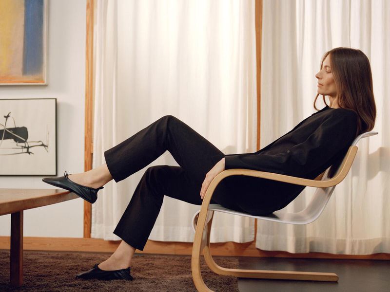 A woman with long brown hair sits in a modern chair, wearing a black blouse and pants, inside a minimalist room with artwork on the walls.