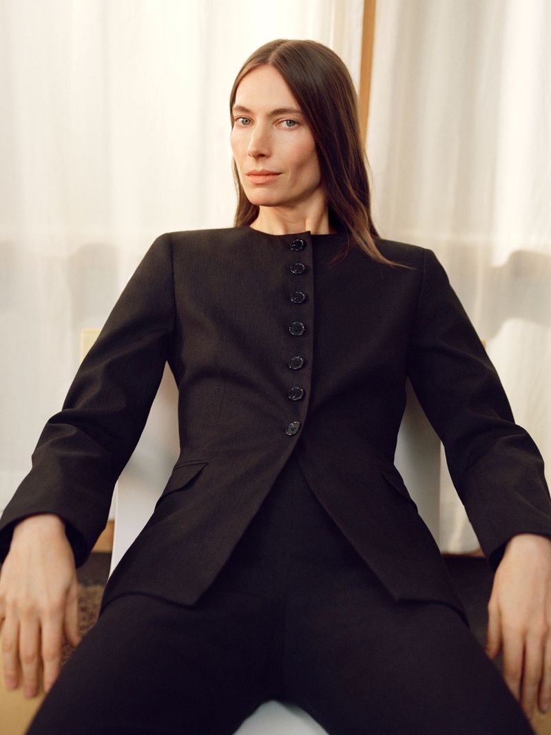 Elegant woman in a black suit poses in front of a neutral background, seated indoors with natural light and a minimalist aesthetic.