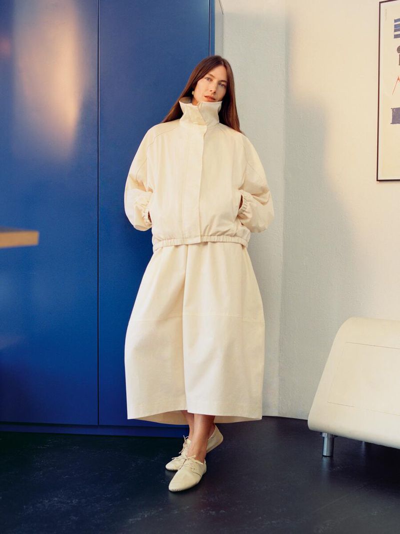 Indoor portrait of a woman in a cream-colored jacket and culottes, standing against a dark blue wall with minimalist decor. Fashion editorial.