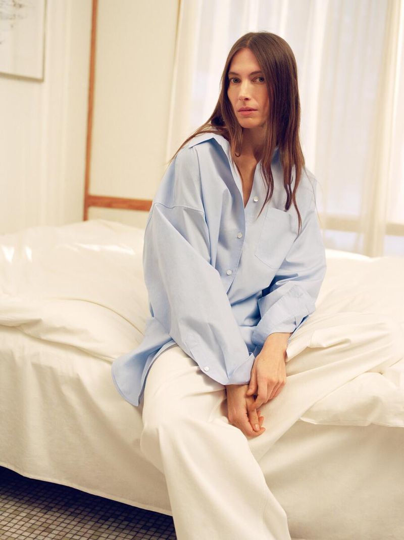 A young woman in a light blue button-down shirt and white pants sits on a bed in a brightly lit indoor setting.