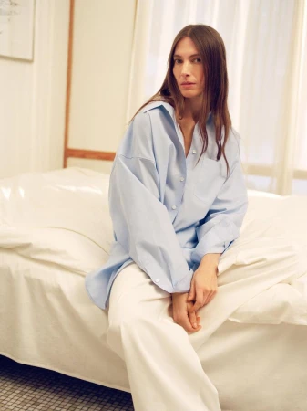 A young woman in a light blue button-down shirt and white pants sits on a bed in a brightly lit indoor setting.