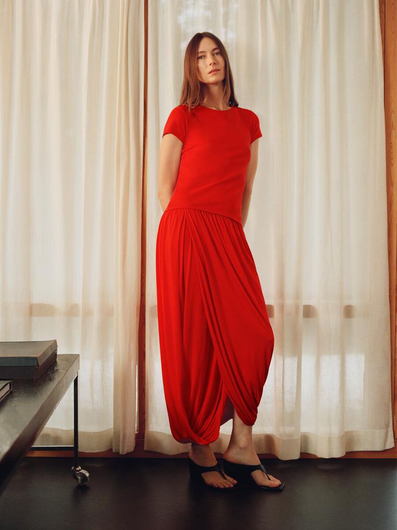 A slender adult woman poses indoors wearing a red top and skirt, along with black sandals. A table and window are visible in the background.