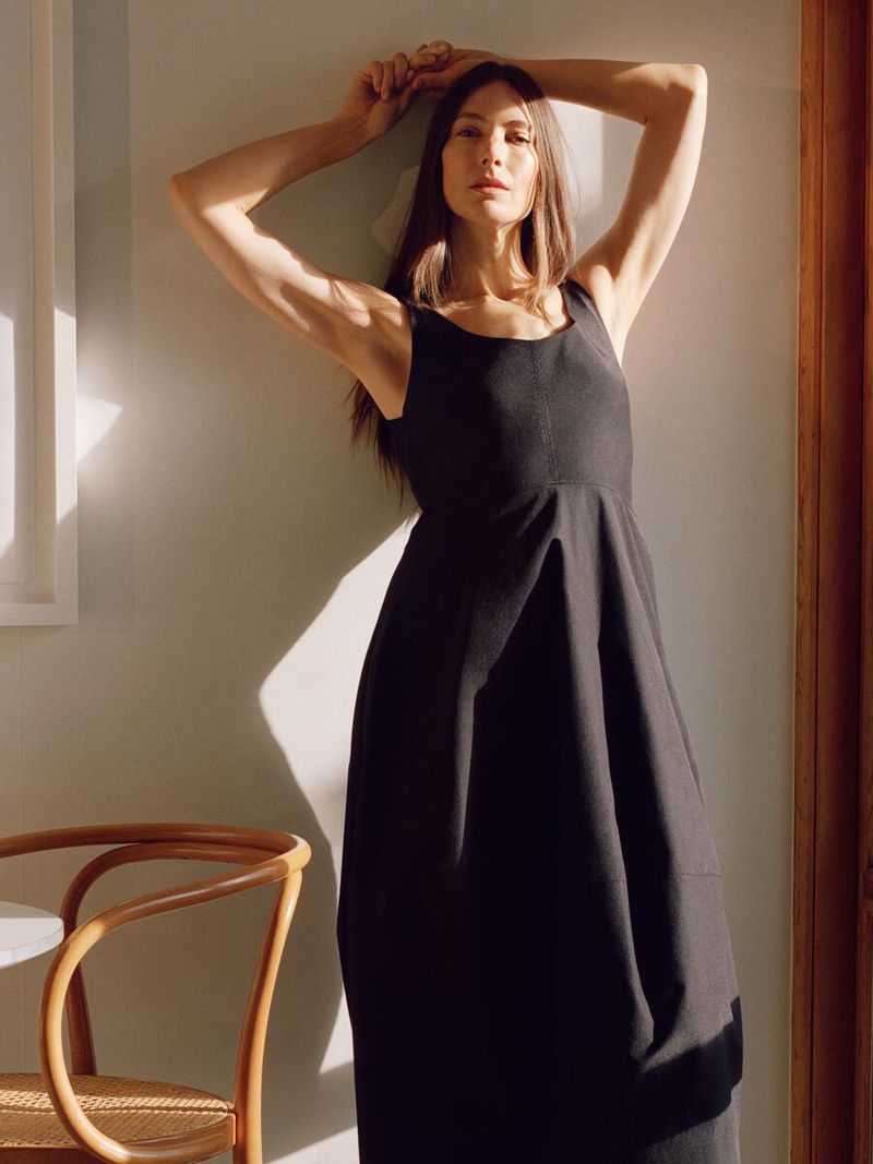A woman in a dark dress leans against a wall in an indoor setting with warm lighting and a modern chair in the frame.
