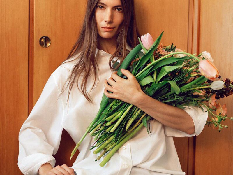 Adult woman with long brown hair in a white shirt holding a flower bouquet in front of a wood paneled wall indoors.