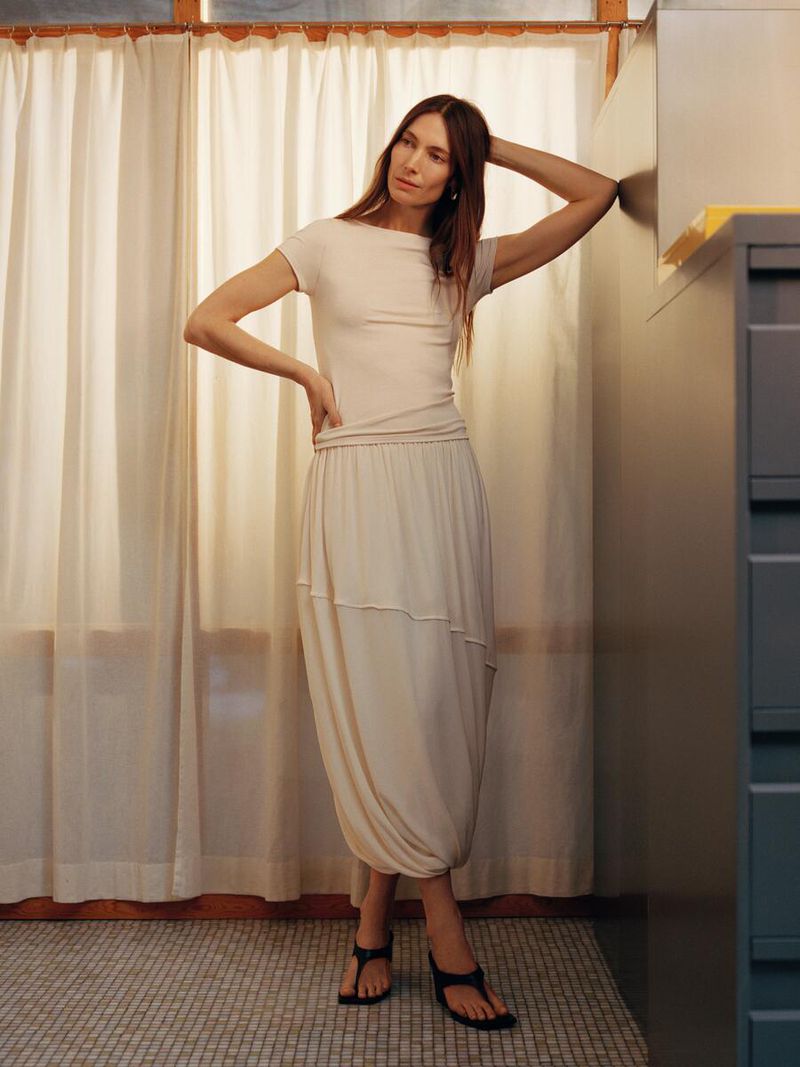 Woman in a neutral-tone dress poses indoors next to a cabinet. She rests one hand on her head and the other on her hip.