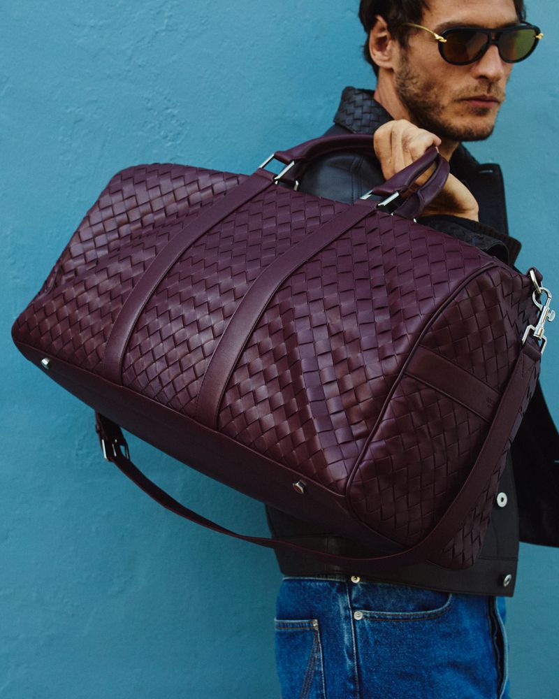 Leather duffel bag with woven pattern, carried by a man in sunglasses, jeans, and leather jacket against a blue wall.