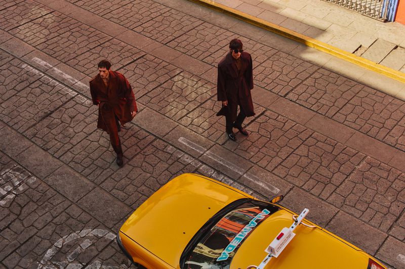 High-angle shot of two men in coats walking on a cobblestone street with a yellow taxi cab in the foreground.