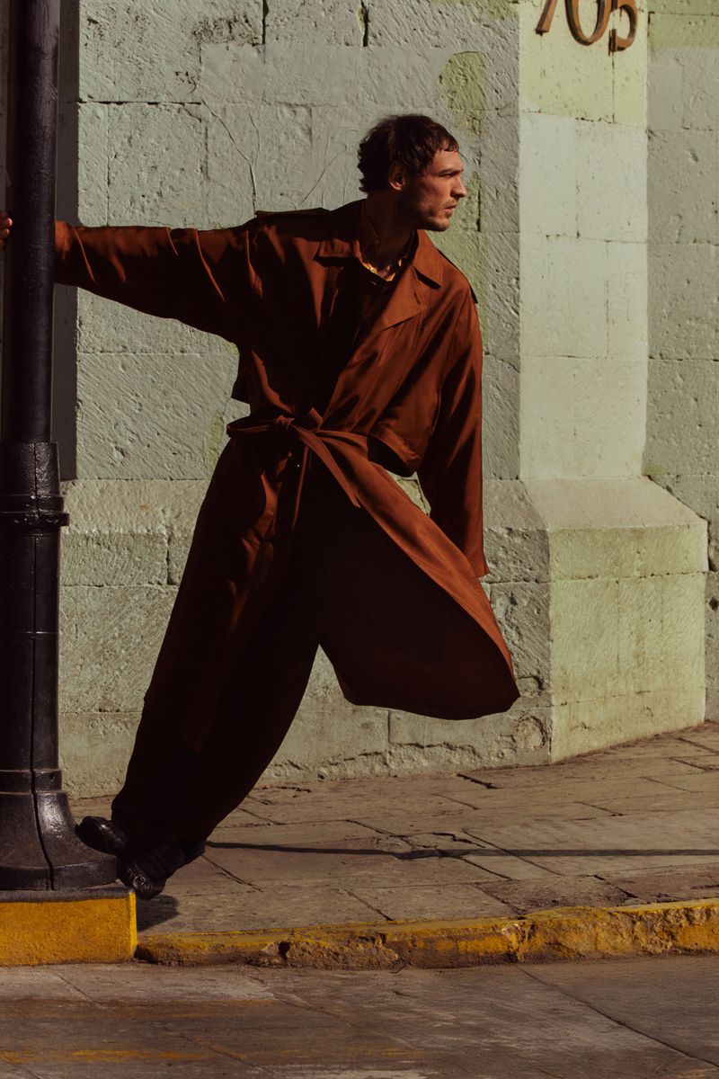 A stylish male model poses against a wall in a brown trench coat and pants, leaning on a black lamppost. Fashion editorial in an urban setting.