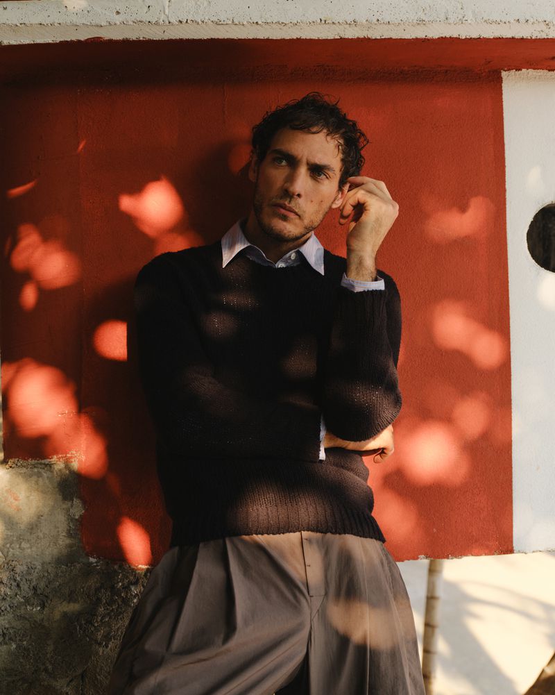 A man leans against a red wall wearing a navy sweater and gray trousers in an indoor studio shot with diffused, natural lighting.
