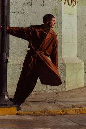 Man wearing a brown trench coat leaning against a dark pole on a stone street background.