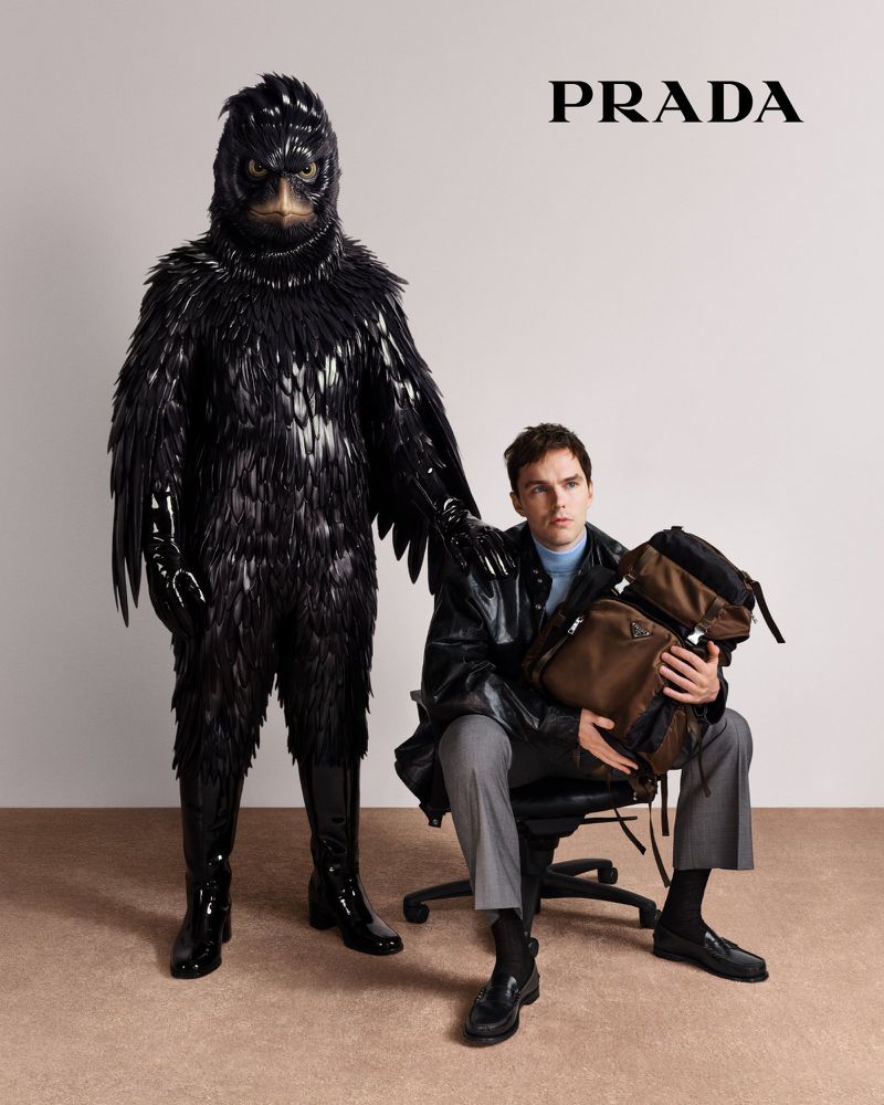 Fashion shot with a human in a black feather costume and a man in a black jacket holding a brown backpack, both sitting indoors.