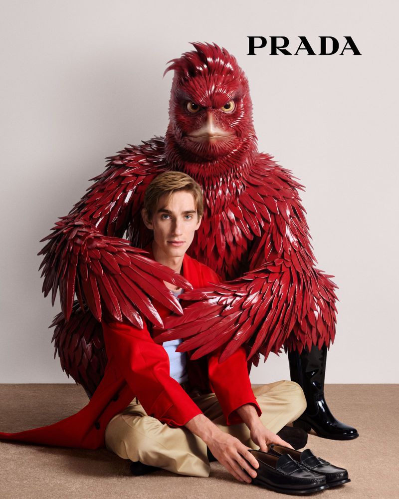 A man in a red jacket sits next to a large bird creature covered in red feathers in a fashion editorial for Prada, indoors.