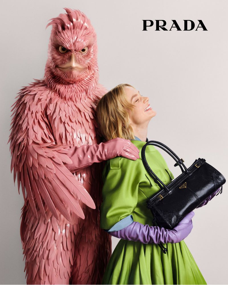 Woman smiles and poses with a black purse next to a person in a pink feathered creature costume in a studio fashion shot.
