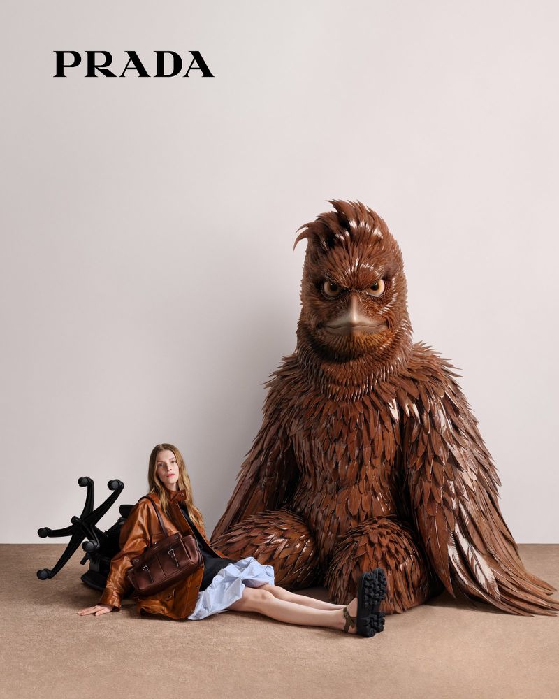 Fashion editorial image featuring a woman in a brown jacket and blue dress sitting next to a large wooden bird sculpture in studio.