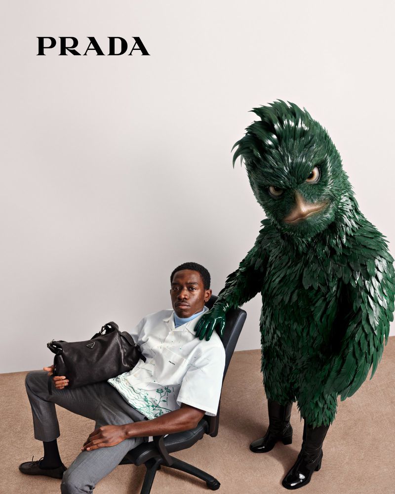 Fashion shot features an adult male model in an office chair next to a person in a green feathered costume and the Prada logo.