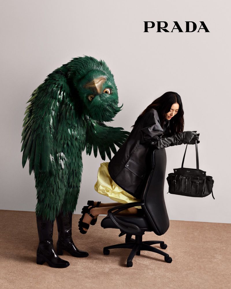 A person in a black jacket and heels poses beside a person in a large green avian costume in a studio for a Prada fashion shot.