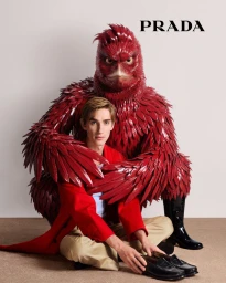 Man sitting cross-legged wearing a red jacket and loafers, embraced by a large red feathery creature with yellow eyes against a white wall.