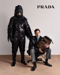 Nicholas Hoult seated beside a towering figure in a detailed black bird costume for a Prada campaign against a neutral background.